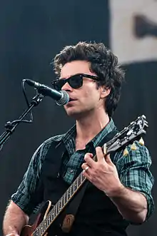 A man wearing sunglasses, a waistcoat and a checked shirt, singing into a microphone while playing an electric guitar