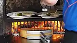 A half raclette cheese being melted on a wood fire while the other half is being scraped from its melted part in a plate.