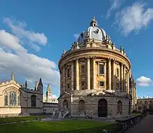 Photo of the Radcliffe Camera from street level showing the Rotunda shaped library with grey top and cream bricks. The railings around the outside have bicycles against them.