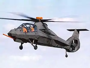 Brown helicopter with faceted surfaces flying. The two crew members inside cockpit are wearing bright orange vests