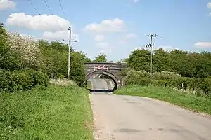 Blue brick single arch bridge carrying former railway over the road into the village.  The first houses are just visible through the arch.  The road surface has been lowered to pass under the bridge.  On either side steep embankments carry the former line, completely overgrown with trees and shrubs