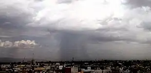 Dark storm clouds under which a grey sheet of rain is falling on a city.