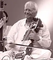 An old man sits cross-legged on a platform and plays a bowed instrument.