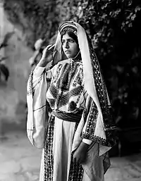 Image 5A young woman from Ramallah, c. 1898-1914.  Until the 1940s, women of Palestine wore elaborate handcrafted garments.  The creation and maintenance of these items played a significant role in their lives.  A knowledgeable observer could determine a woman's village of origin and social status from her clothing.  The circular band near this woman's forehead is a ring of coins made from a portion of her dowry money, and indicates that she is unmarried.'Ali Ra'if Efendi, (edited by Durova)