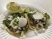 Photograph of two tacos and a small container of salsa on a paper plate