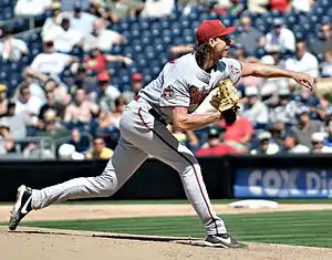 A man wearing a lightly colored baseball uniform and a red cap throws a pitch.