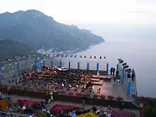 An orchestra before a performance on an outdoor stage on a balcony with water in the background.