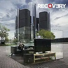 The cover image features skyscrapers in center under cloudy blue sky. Above which, on the ground, transparent glass-type cuboidal body is placed. Inside which, appears a living room with Eminem seated on sofa watching television. On top-left corner, in bold and capitalised format, the title RECOVERY appears.