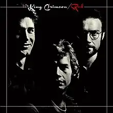 A black-and-white photograph of three men facing the camera. The words "King Crimson / Red" are written at the top-centre of the photograph.