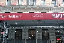 The facade of the base in 2023. The second floor is covered by a red scaffolding reading "The Redbury" and "Marta".