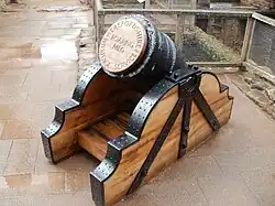 A squat black mortar, the end gapped with a wooden plug on which is carved "Roaring Meg"; the mortar has wooden supports with black metal brackets.