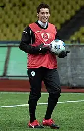 A dark-haired white man wearing a training bib, holding a ball and smiling.