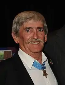 Head and shoulders of a white man with a pointed mustache, wearing a star-shaped medal on a blue ribbon around his neck.