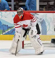 An ice hockey goaltender wearing a red mask, white pads and a white and red jersey with a maple leaf logo. He is bent over with his hands at his knees and his head looking forward.