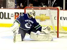 An ice hockey goaltender with his right leg and glove stretched out to make a save. He wears a blue jersey, white pads and a white helmet.