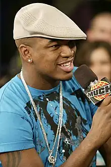 A man holds a microphone while he stands in front of a crowd. He smiles and wears a blue T-shirt with a dark stamping of man on it, and a light brown beret.