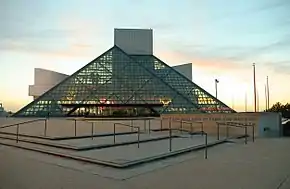 An extended glass pyramid in front of a tall gray building with strangely shaped outcroppings