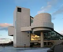 A gray tiled building rises over a lake, with a cylinder set on a narrow pole, and a sloping glass wall on one end.