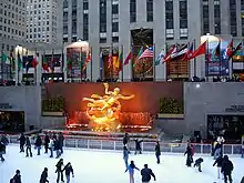 Ice skaters on a rink below a golden sculpture and a row of national flags that fly in front of a stone tower.