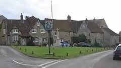 Street scene. Triangular area of grass with village sign on wooden post and stone cross behind. Stone houses with tiled roofs n the background.