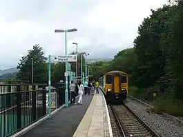 View along country station platform with bilingual exit signage. Passengers wait to board a two carriage train, which is stationary on the single track. Platform, train and track curve slightly right to left. Trees in full leaf stand alongside the track, with mountains in the distance.