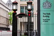 The entrance to the Rolls Building. A pair of black iron gates with large lanterns on top, next to a large glass sign saying, "The Rolls Building".