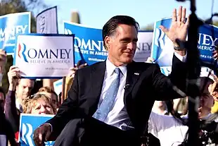 Mitt Romney sitting outdoors during daytime, with crowd behind him holding up blue and white "Romney" signs