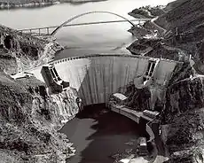 The downstream face of renovated Roosevelt Dam, with a concrete face. A bridge spanning Lake Roosevelt is seen in the background.