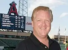 A man is smiling at the camera with the backdrop consisting of the interior of a baseball stadium with the scoreboard displayed prominently.