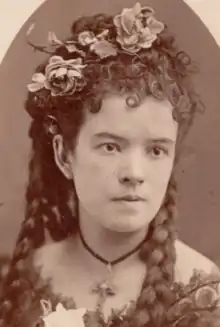 A young white woman with her hair curled, braided, and decorated with flowers; she is wearing a large cross on a velvet band at her neck; her dress is bare at the shoulders, and embellished with lace.