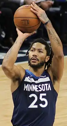 Hip length view of man raising the basketball to shoot free throw, with black moustache, beard, and short dreadlocks, thin white headband, wearing navy blue Timberwolves uniform