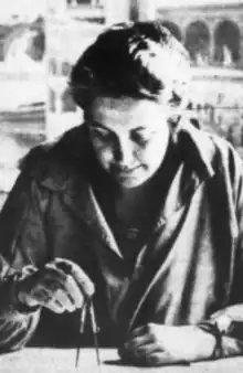 A white woman seated at a table, holding a protractor in one hand