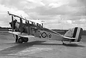 Manned single-engined military biplane parked on airfield, with propeller spinning