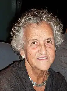 Portrait photograph of smiling, gray-haired woman wearing a brown V-neck shirt and a silver necklace.