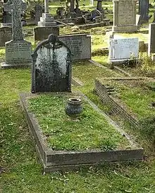 A granite headstone almost completely covered in black moss, in a grassy cemetery