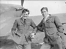 A black and white photograph of two smiling men in uniform standing in front of and partly leaning on the wing of a fighter aircraft