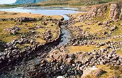 A stone lined ditch of primitive construction leads from a small lake. Rocky heathland lies on either side and there are tall cliffs in the distance.