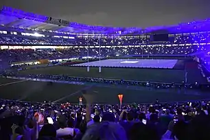 A rugby field with drummers on the perimeter, and a large multicoloured flag in the middle.