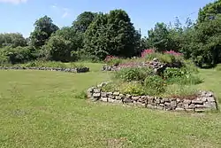 Low stone walls in grass, surrounded by trees with a house in the distance.