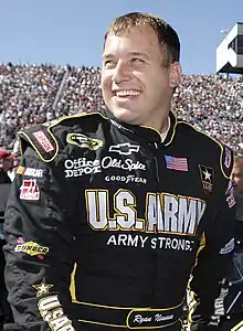 Man in his early 30s, wearing a black racing suit with sponsor logos