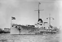 A large warship sits in harbor, with two smaller boats alongside. High-rise buildings are seen in the distance.