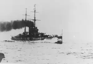 A large battleship steams through the water. Water breaks against the bow as heavy dark smoke emerges from the ship's two funnels.