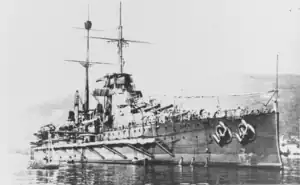 A large battleship sits still in the water near land. Several small boats can be seen in the foreground while a large hill appears behind the battleship in the background.