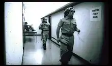 Black and white photo of a white hallway with a plaque reading "Stanford County Prison". Two white male guards in uniforms wearing sunglasses walk along the hallway. One white male guard is sitting on top of a table in a corner in the background.