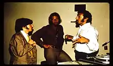 Photo of three white male people wearing shirts and ties talking to each other in front of a white wall. They are standíng beside a tape recording machine.