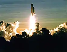 A space shuttle launches into a dawn sky. Clouds in the sky, in the launch plume and from the flame trench, are visible, as is the scaffolding-like launchpad and some vegetation silhouetted in the foreground.