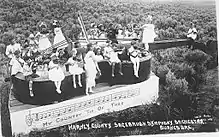 Black and white image of a group of children holding stringed instruments, sitting and standing in various position on a violin-shaped float in an open field.