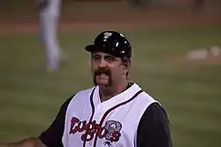 A smiling, mustachioed man in a white-and-black baseball jersey and black baseball helmet