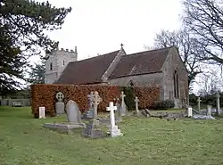 Gray stone building with red tiled roof, partially obscured by a hedge. A square tower is at the far end. The foreground includes several crosses and gravestones.