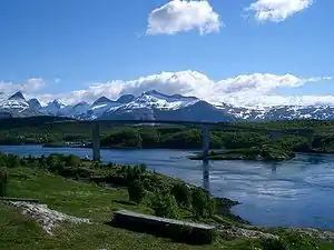 A cantilever bridge crosses a sound surrounded by green forests and jagged mountains in the backdrop